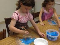 Two girls, both wearing pink, use paintbrushes to glue blue tissue paper onto an art project.